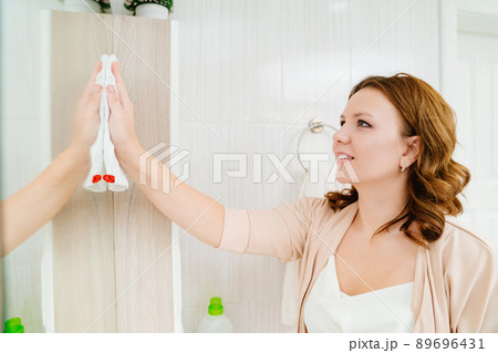 an attractive housewife washes the bathroom mirror with a special glass cleaner. an attractive housewife washes the bathroom mirror with a special glass cleaner. 89696431