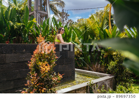 Surrounded by palm trees, a bunk pool on the upper tier stands a bather in a bright top and sunglasses from the sun Surrounded by palm trees, a bunk pool on the upper tier stands a bather in a bright top and sunglasses from the sun 89697350