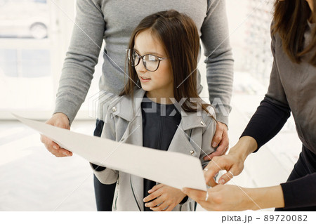 Father and daughter choosing glasses in optics store. Female optometrist helping customers choosing glasses. Little girl in optics store choosing glasses with her father. Father and daughter choosing glasses in optics store. Female optometrist helping customers choosing glasses. Little girl in optics store choosing glasses with her father. 89720082