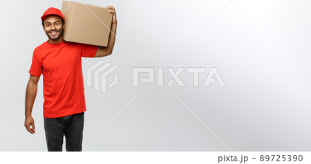 Delivery Concept - Portrait of Happy African American delivery man in red cloth holding a box package. Isolated on Grey studio Background. Copy Space. Delivery Concept - Portrait of Happy African American delivery man in red cloth holding a box package. Isolated on Grey studio Background. Copy Space. 89725390