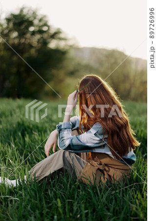 A woman sits on the green grass in a park with her back to the camera and relaxes in nature in the summer sunset evening light A woman sits on the green grass in a park with her back to the camera and relaxes in nature in the summer sunset evening light 89726996