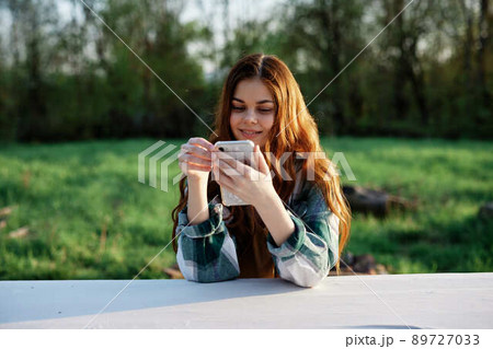 A beautiful woman relaxing and working on her phone sitting in nature in the park among the trees smiling and holding her smartphone in her hand lit by the bright sunset light A beautiful woman relaxing and working on her phone sitting in nature in the park among the trees smiling and holding her smartphone in her hand lit by the bright sunset light 89727033