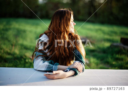 Girl with phone in hand sitting in nature in the park at the table smiling beautifully and looking at the camera with her red hair lit by the sunset sunlight of summer Girl with phone in hand sitting in nature in the park at the table smiling beautifully and looking at the camera with her red hair lit by the sunset sunlight of summer 89727163