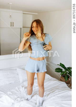Young teen girl in pajama standing on her bed and singing a song. Cheerful caucasian adolescent girl with headphones dancing on her bedroom .  89731456