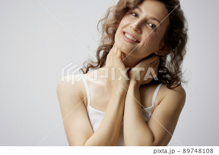 Joyful awesome curly beautiful woman in basic white t-shirt touches neck smiles at camera posing isolated on over white background. People Emotions Lifestyle concept. Copy space 89748108