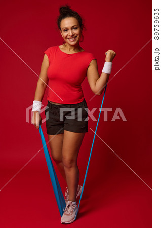 Attractive athletic fit woman doing body weight workout using elastic strap pulling with arms for shoulder training against red background with copy ad space Attractive athletic fit woman doing body weight workout using elastic strap pulling with arms for shoulder training against red background with copy ad space 89759635