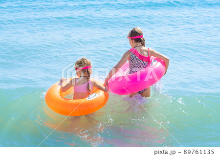 Two beautiful little girls play and have fun in sea water on the beach. Vacation or holiday concept. Children girls with inflatable swimming rings having fun in blue sea water 89761135
