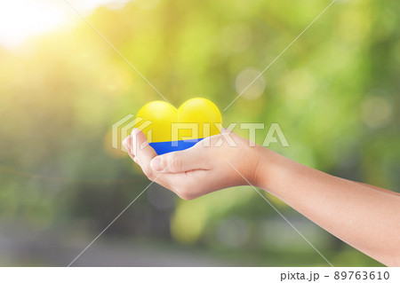 Child hand holding heart in the color flag nation of Ukraine over green nature background. Concept of ending the war in Ukraine. Child hand holding heart in the color flag nation of Ukraine over green nature background. Concept of ending the war in Ukraine. 89763610