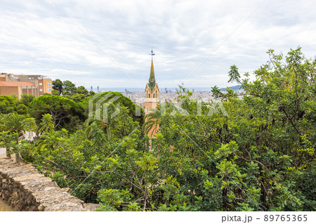 Barcelona, Spain - September 22, 2021: The steeple of the small village in Parc Guell. Park Guell is the famous architectural city art by Antoni Gaudi. Tourist hot spot. Cityscape in the background Barcelona, Spain - September 22, 2021: The steeple of the small village in Parc Guell. Park Guell is the famous architectural city art by Antoni Gaudi. Tourist hot spot. Cityscape in the background 89765365