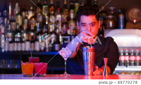 Young bartender mixing ingredients in the shaker with a spoon - a glass with ice - cocktails on the stand 89770278