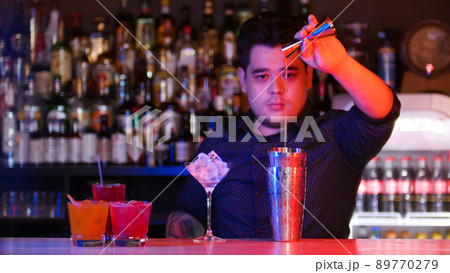 Young bartender standing in front of his bartender devices - pouring a syrup in the shaker - cocktails on the stand 89770279