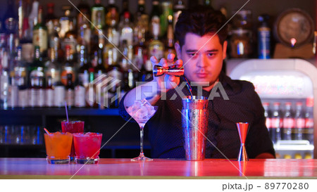 Young bartender standing in front of his bartender devices - pouring a syrup in the shaker - a glass with ice - cocktails on the stand 89770280