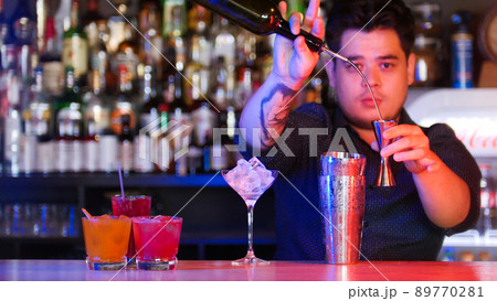 A man artender standing in front of his bartender devices - pouring a syrup in the measurer - a glass with ice - cocktails on the stand 89770281