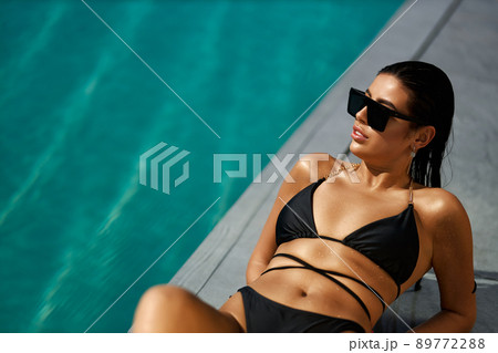 Tanned girl in black bikini laying on grey pool deck and looking away against turquoise water surface. Above view of wet lady in sunglasses sunbathing near pool on vacation. Concept of relaxation. 89772288