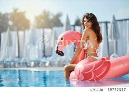 Smiling brunette female sitting on coral inflatable flamingo, while floating in pool. View from shoulder of attractive girl in bikini having fun in pool against sunlight outdoors. Concept of leisure. 89772357