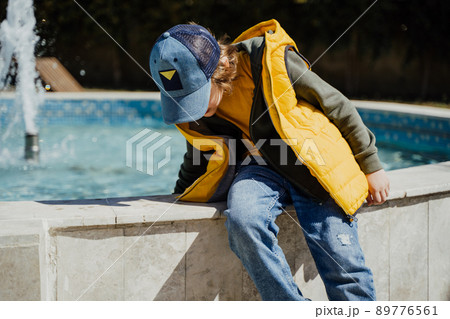 Schoolboy sitting on a fountain in public park during spring sunny day playing with water. Kid in yellow vest chilling in park with water sprinkling in the background. Summer holidays outside 89776561