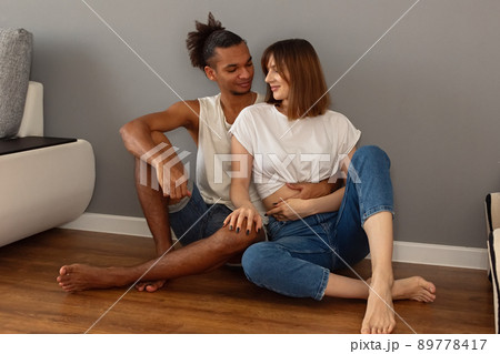 A couple, a guy and a girl are sitting at home on the floor against a gray wall 89778417