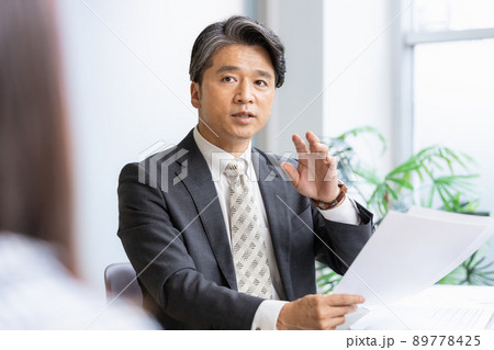 書類を見ながら手前の人物と話をするビジネスマン 89778425
