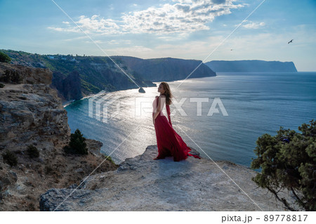 A girl with flowing hair in a long red dress stands on a rock above the sea. The stone can be seen in the sea. Sunny path to the sea from the sun. A girl with flowing hair in a long red dress stands on a rock above the sea. The stone can be seen in the sea. Sunny path to the sea from the sun. 89778817