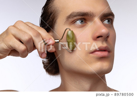 Young attractive man with perfect smooth skin, applying face massagers isolated over white studio background. Cosmetics, health care, skin care, beauty 89780500