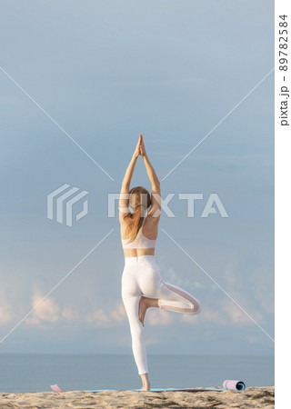 Girl in white sporty clothes practicing yoga, tree pose on the beach. Vrikshasana. Young woman doing yoga with the sea behind. 89782584