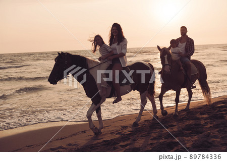The family spends time with their children while riding horses together on a beautiful sandy beach on sunet.  89784336