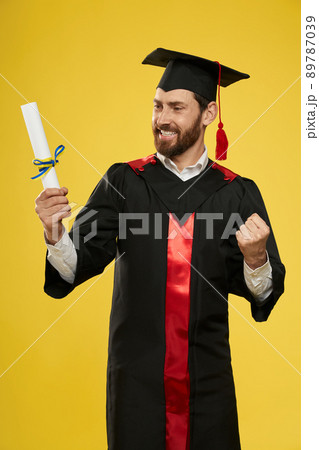 Front view of happy student with mortarboard holding diploma. Boy with beard graduating from college, university, cheerful, successful. Isolated on yellow studio background. Front view of happy student with mortarboard holding diploma. Boy with beard graduating from college, university, cheerful, successful. Isolated on yellow studio background. 89787039