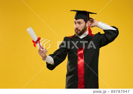 Front view of student standing, looking at diploma, mixed up, surprised. Young male wearing mortarboard and graduate gown graduating from college. Isolated on yellow background. 89787047