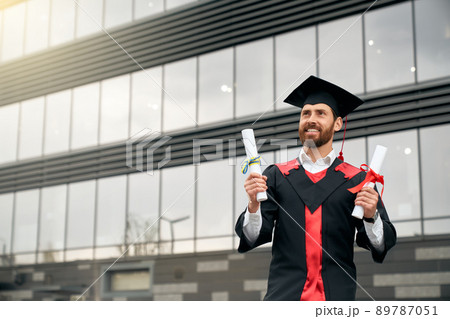 Front view of student in mortarboard and graduate gown holding two diplomas. Handsome young man graduating from college, smiling, happy, cheerful. Concept of education and youth. Front view of student in mortarboard and graduate gown holding two diplomas. Handsome young man graduating from college, smiling, happy, cheerful. Concept of education and youth. 89787051