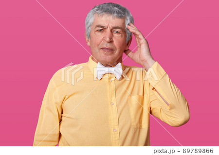 Studio shot of handsome old man keeps hand on temple, has headache, wears yellow shirt and white bow tie, has embrassed facial expression, isolated over pink studio background. People and age concept. 89789866