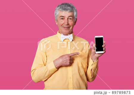 Portrait of attractive mature man with gray hair and wrinkles, dressed yellow shirt in one tone and white bow tie, holding phone with blank screen, points with fore finger on copy space for your text. Portrait of attractive mature man with gray hair and wrinkles, dressed yellow shirt in one tone and white bow tie, holding phone with blank screen, points with fore finger on copy space for your text. 89789873