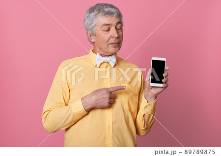 Close up portrait of senior man wearing yellow shirt with white bowtie showing black blank smart phone screen, looking at his device, posing over pink background, looks serious and concentrated. Close up portrait of senior man wearing yellow shirt with white bowtie showing black blank smart phone screen, looking at his device, posing over pink background, looks serious and concentrated. 89789875