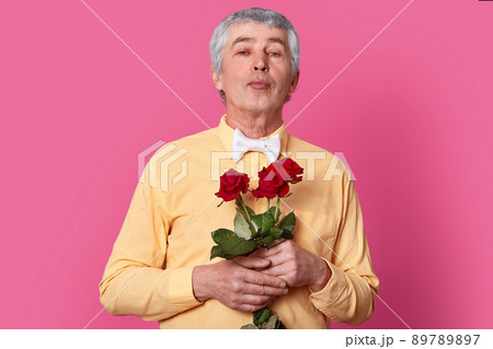 Isolated shot of attractive wrinkled grey haired man holds red roses, dressed in yellow elegant shirt and bowtie, keeps lips folded, going to kiss someone. Grandfather recieves flowers on birthday 89789897