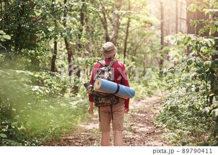 Back view of male hiker with backpack and sleepin pad on his back, walking in forest, being surrounded with green trees, wearing casual comfortable clothing. Adventure and traveling concept. Back view of male hiker with backpack and sleepin pad on his back, walking in forest, being surrounded with green trees, wearing casual comfortable clothing. Adventure and traveling concept. 89790411