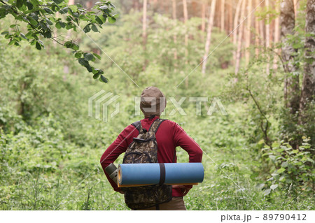 Hiking. Man wearing red sweater and cap in hike, having backpack and sleeping pad, looking towards. Sporty man hiking on green trees background. Rear view. Hiking, tourism, healthy lifeslyle concept. 89790412