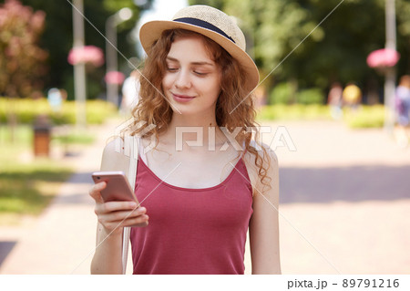 Delighted happy young lady having pleasant facial expression, smiling sincerely, reading news, holding smartphone, using it actively, wearing red shirt and fashionable summer hat. Social medias. Delighted happy young lady having pleasant facial expression, smiling sincerely, reading news, holding smartphone, using it actively, wearing red shirt and fashionable summer hat. Social medias. 89791216