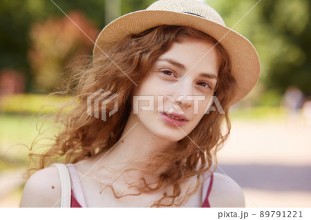 Close up portrait of attractive young lady in straw hat. Foxy haired charming woman with brown eyes looking at camera with gentle, has calm facial expression, posing isolated over park background. 89791221