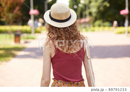Back view of slender curly haired female wearing straw hat and red shirt, having bag on her shoulder, walking along paved street, spending time around nature in local park. Summer day concept. Back view of slender curly haired female wearing straw hat and red shirt, having bag on her shoulder, walking along paved street, spending time around nature in local park. Summer day concept. 89791233
