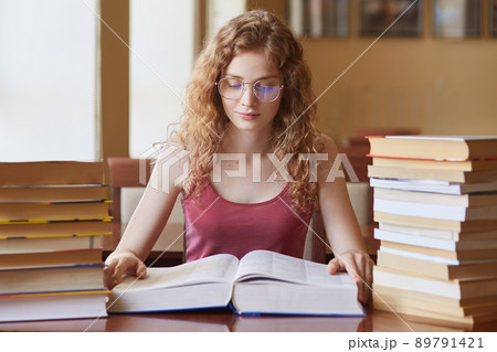 Portrait of hard working thoughtful college student with fair curly hair reading huge book, doing research, preparing for her lessons, looking attentively, having many tasks to do. Student life. Portrait of hard working thoughtful college student with fair curly hair reading huge book, doing research, preparing for her lessons, looking attentively, having many tasks to do. Student life. 89791421