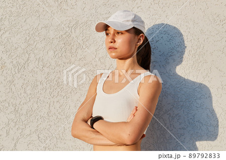 Portrait of serious confident female wearing white top and visor cap, standing isolated over gray wall outdoor with folded hands, looking in distance, sporty woman after work out. Portrait of serious confident female wearing white top and visor cap, standing isolated over gray wall outdoor with folded hands, looking in distance, sporty woman after work out. 89792833