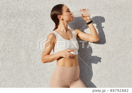 Outdoor shot of beautiful female runner standing outdoors near gray wall drinking water from bottle, fitness woman taking a break after workout, healthy lifestyle. Outdoor shot of beautiful female runner standing outdoors near gray wall drinking water from bottle, fitness woman taking a break after workout, healthy lifestyle. 89792882