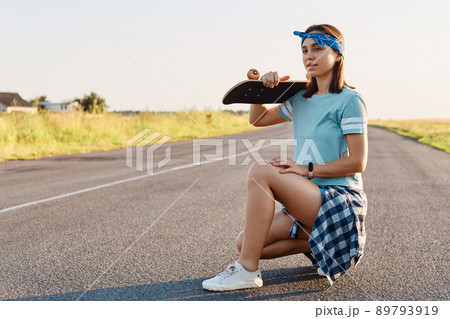 Young adult attractive girl with dark hair posing with a longboard over shoulders while sitting on the road in summer, looking away with pensive expression. Young adult attractive girl with dark hair posing with a longboard over shoulders while sitting on the road in summer, looking away with pensive expression. 89793919