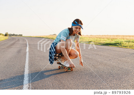 Portrait of happy beautiful dark haired female wearing casual attire and hair band surfing sitting on skateboard, having fun outdoor alone, healthy active lifestyle. 89793926