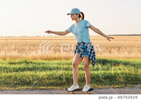 Side view of slim sporty female wearing casual style t shirt and visor cap skateboarding on street on asphalt road, happy active pastime in summer outdoor. 89793929