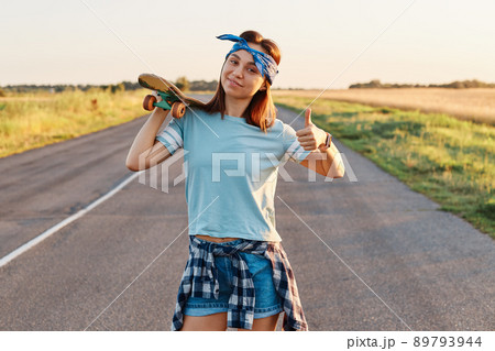 Cheerful girl with stylish hairband posing with a longboard over shoulders while walking on the road, looking at camera with happy emotions and showing thumb up. Cheerful girl with stylish hairband posing with a longboard over shoulders while walking on the road, looking at camera with happy emotions and showing thumb up. 89793944