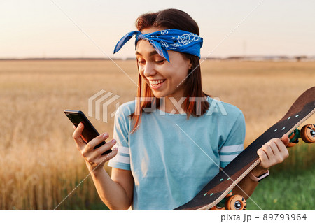 Smiling female with dark hair wearing t shit and hair band holding in hands skateboard and smart phone, looking at cell phone display with happiness, reading excellent news from message. 89793964