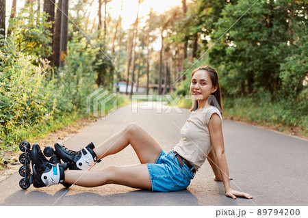 Young adult beautiful woman with dark hair wearing beige t shirt and jeans short, sitting on road in summer park, having rest while rollerblading, healthy lifestyle. 89794200