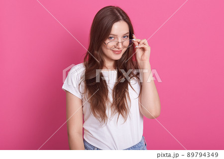 Indoor shot of attractive young woman with long hair keeps hand on rim of spectacles, wearing casual outfit, model posing against pink studio wall, looks smiling directly at camera. People concept. Indoor shot of attractive young woman with long hair keeps hand on rim of spectacles, wearing casual outfit, model posing against pink studio wall, looks smiling directly at camera. People concept. 89794349