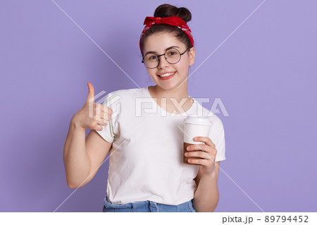 Happy young woman showing thumb up and holding take away coffee, looking smiling directly at camera, wearing casual attire and red hairband, being in good mood. Happy young woman showing thumb up and holding take away coffee, looking smiling directly at camera, wearing casual attire and red hairband, being in good mood. 89794452