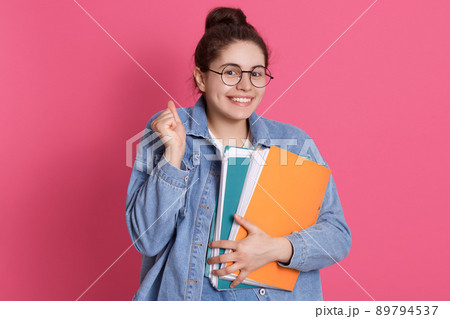 Young smiling student in eyeglasses and denim jacket standing with paper folders isolated over rosy background, clenching fist, looking smiling at camera. 89794537